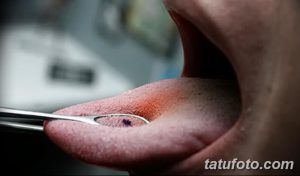 Фото тату: Крупный план языка, зажатого медицинским инструментом во время процедуры пирсинга. Видна подготовка к введению украшения.