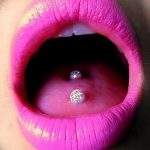 Фото пирсинга языка с серебристым украшением, украшенным кристаллами. Язык высунут, демонстрируя пирсинг и ярко-розовые губы. Гламурный и привлекательный образ.