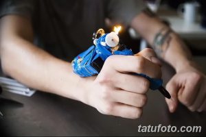 Руки держат самодельную тату-машинку, из которой идет дым. На руке мастера видны татуировки. Фотография демонстрирует процесс нанесения татуировки.
