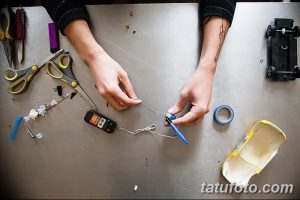Руки мастера собирают самодельную тату-машинку из различных деталей. Вид сверху, на рабочем столе разложены инструменты.
