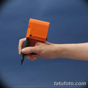 Фотография оранжевой тату-машинки, удерживаемой в руке. Изображение демонстрирует простоту и доступность устройства.