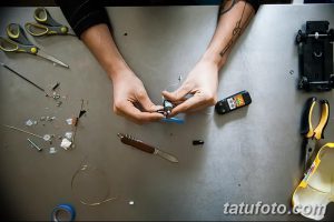 Фотография рук, работающих с мелкими деталями тату-машинки на сером столе. Видны инструменты, провода и детали, создающие атмосферу мастерской.
