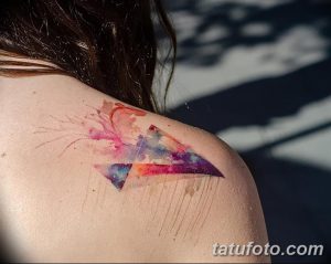 Фото тату акварельной стрелы на плече. Нежные переходы цветов, брызги краски, легкий и воздушный дизайн.