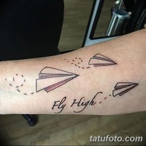 Фото татуировки на предплечье с изображением нескольких бумажных самолетиков и надписью 'Fly High'. Тату выполнена в коричневых тонах с пунктирным следом.