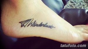 Фото тату: небольшой рисунок бумажного самолетика и надпись 'Wanderlust' на стопе. Черно-серые тона, минималистичный стиль.