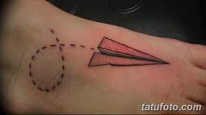 Фото тату: цветная татуировка бумажного самолетика на стопе, с красными акцентами и пунктирной линией, символизирующей полет.