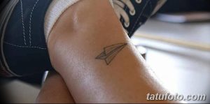 Фото тату: крошечный бумажный самолетик, выполненный в серо-коричневых тонах, расположен на лодыжке.