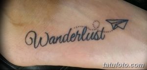 Фото татуировки на стопе с надписью "Wanderlust" и изображением бумажного самолетика. Стильный рисунок тату для любителей путешествий.