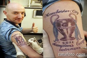Фото тату кубка Manchester City на плече фаната. Рисунок выполнен в стиле контурной графики с добавлением красных акцентов. Изображение сопровождается надписью и годом победы.