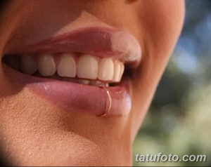 Фотография демонстрирует пирсинг нижней губы с использованием небольшого серебристого кольца. Пирсинг расположен сбоку от центра губы.