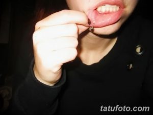 Фото тату: девушка держит губу, демонстрируя свежий пирсинг с небольшим металлическим кольцом. Видно место прокола и начальный этап заживления.