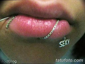 Крупный план губы с проколом, украшенным цепью и подвеской с надписью "SEXY". Фото демонстрирует провокационный и привлекательный образ.