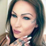 Фотография привлекательной девушки с татуировками на руках и лице. Татуировка на лице представляет собой надпись.