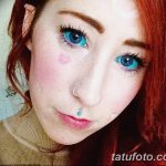 Фотография девушки с рыжими волосами и татуировкой в виде сердца на щеке. На лице заметен пирсинг. Изображение демонстрирует необычный стиль.