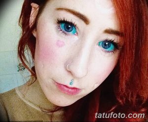 Фотография девушки с рыжими волосами и татуировкой в виде сердца на щеке. На лице заметен пирсинг. Изображение демонстрирует необычный стиль.