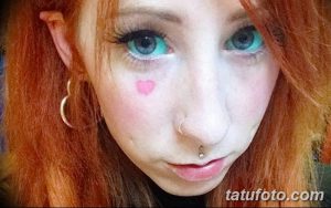Фото тату на лице девушки: маленькое розовое сердце на щеке, рыжие волосы, голубые глаза. Нежные идеи для тату.