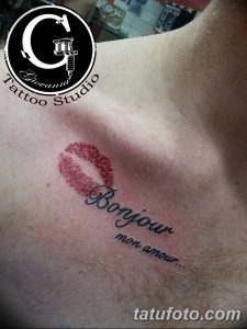 Фото тату: отпечаток губ и надпись 'Bonjour mon amour...' на груди, выполненные в красных и черных тонах.