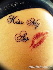 Фото тату на бедре: красные отпечатки губ и надпись 'Kiss My Ass' выполнены в каллиграфическом стиле. Выражает бунтарский характер и независимость.