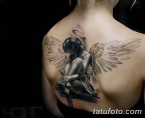 Фото татуировки ангела с опущенной головой на лопатке. Черно-белый рисунок, реалистичный стиль, детализированные крылья, печальное выражение лица.