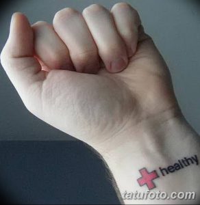 Фото тату на запястье: красный медицинский крест и надпись 'healthy'. Простая татуировка, символизирующая здоровье.