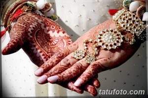 Фото тату мехенди на руках невесты, покрытых сложными узорами в красных тонах. Руки украшены браслетами и кольцами.
