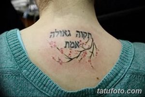 Фото тату: надпись на иврите и изображение цветущей ветки, выполненные черными и красными чернилами на затылке. Текст расположен над веткой, создавая гармоничный и романтичный образ.