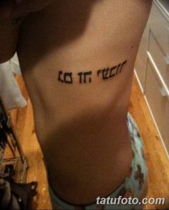 Фото тату: Черная татуировка с ивритским текстом, изогнутым вдоль линии тела на боку. Элегантный и женственный дизайн.