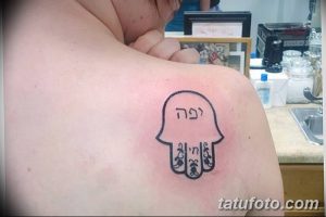 Фото тату Хамса на плече, выполненное в минималистичном стиле с добавлением надписи на иврите. Небольшой размер и четкие линии.
