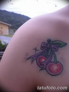 Фото татуировки вишни, украшенной фиолетовым бантом. Нежный стиль, пастельные тона, детализированная прорисовка.