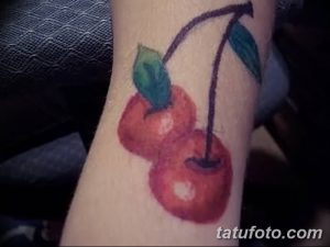 Фото тату двух красных вишен с зелеными листьями на руке. Нежный и романтичный рисунок татуировки, выполненный в технике акварели.