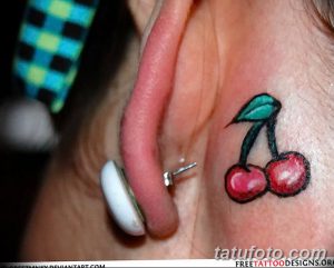 Фото маленькой татуировки вишни за ухом. Красные ягоды и зеленый лист создают изящный и привлекательный эскиз.