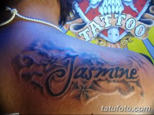 Фото тату: Татуировка с именем "Jasmine" и цветами на женской лопатке, выполненная в реалистичном стиле с использованием черных и серых оттенков.