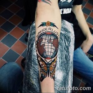 Фото тату в стиле традишнл с изображением Земли, бабочки и надписью 'Wanderlust' на предплечье. Яркие цвета и четкие линии.