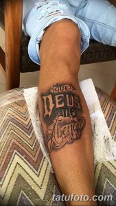 Фото татуировки на голени: надпись "Que Deus te abencoe" (Пусть Бог благословит тебя) выполненная в каллиграфическом стиле, с декоративными элементами. Черно-серая татуировка.