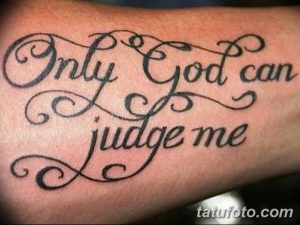 Фото тату надписи 'Only God can judge me' с декоративными завитушками на предплечье. Черный цвет, каллиграфия, религиозный смысл.