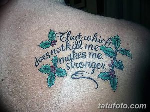 Фото тату надписи 'That which does not kill me makes me stronger' с ветками и ягодами на плече. Цветная татуировка, растительный орнамент, вдохновляющая фраза.