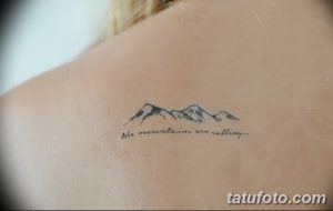 Фото минималистичной татуировки с изображением гор и надписью 'the mountains are calling...' на лопатке девушки.