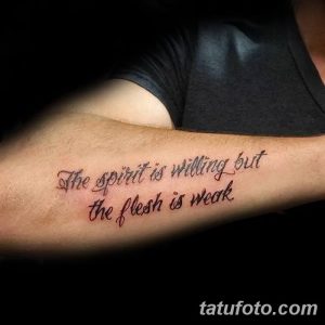 Фото тату с надписью 'The spirit is willing but the flesh is weak' на предплечье мужчины. Черные чернила, каллиграфический шрифт.