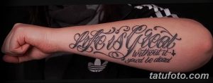 Фото тату: стильная надпись 'Life is great without it you'd be dead' с декоративными элементами на предплечье мужчины. Черно-белая татуировка, выражающая любовь к жизни.