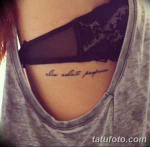 Фото тату: черная надпись на латыни 'alia volat propriis' расположена под грудью, создавая элегантный и женственный образ. Идеально для любителей минимализма.