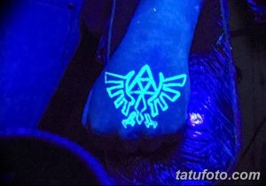 Фотография татуировки с символом из игры The Legend of Zelda, выполненной на стопе и светящейся в ультрафиолете. Минималистичный и узнаваемый дизайн.