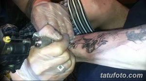 Фото тату в процессе нанесения: рука мастера с тату-машинкой наносит рисунок на руку клиента. Видны перчатки и контуры рисунка.