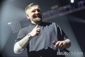 Фотография мужчины на сцене с микрофоном в руке и татуировками на предплечье. Мужчина улыбается и показывает большой палец вверх.