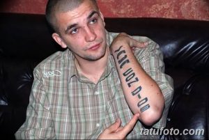 Фотография мужчины в клетчатой рубашке с татуировкой на предплечье, смотрящего в камеру. Татуировка представляет собой надпись.