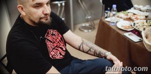 Фотография мужчины в черной футболке с татуировками на руке, сидящего за столом с едой. Мужчина выглядит расслабленным и непринужденным.