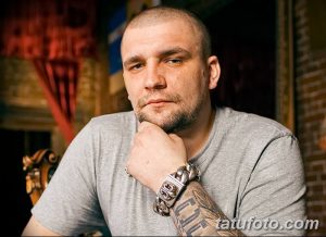 Фотография мужчины с короткой стрижкой и татуировками на предплечье, сидящего и задумчиво смотрящего в камеру. Татуировки выполнены в темных тонах.