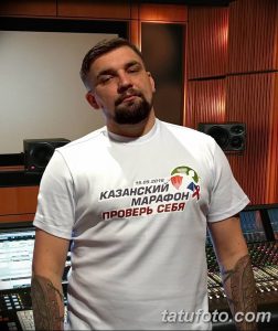 Баста в звукозаписывающей студии, демонстрируя татуировки на предплечье. На нем белая футболка.