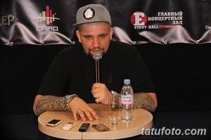 Баста сидит за столом на пресс-конференции, демонстрируя татуировки на предплечье. На голове - кепка.