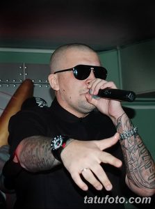 Мужчина с татуировками на руке, выступающий с микрофоном и в темных очках. Фотография передает атмосферу уверенности и стиля.