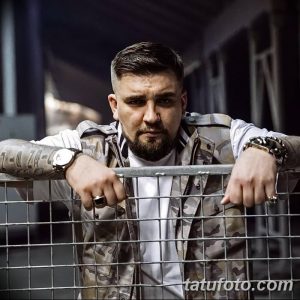 Мужчина с татуировками на руках, стоящий у металлической ограды. Фотография передает атмосферу уверенности и стиля.